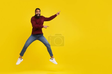 Téléchargez les photos : Émotionnel excité millénial barbu oriental gars sautant dans les airs et pointant vers vide espace maquette, montrant la publicité, isolé sur fond de studio jaune, pleine longueur - en image libre de droit