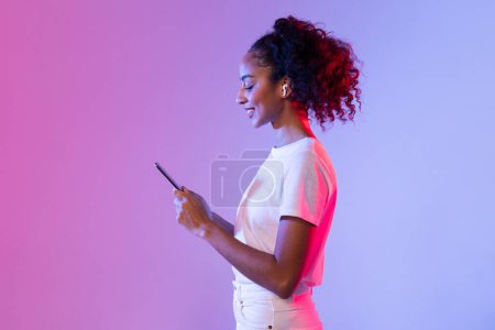 Photo for Profile view of joyful black lady with curly hair using smartphone and wireless earbuds, set against gradient of pink to purple hues, neon background - Royalty Free Image