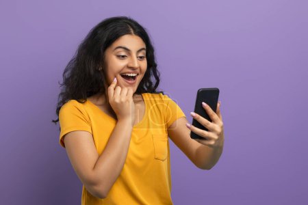 Überglückliche junge Frau untersucht ihren Smartphone-Bildschirm mit einer ekstatischen Reaktion auf violettem Hintergrund