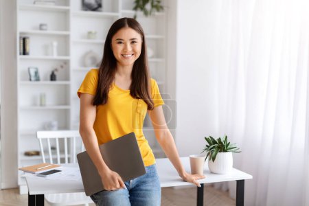 Téléchargez les photos : Souriante jeune femme asiatique tenant ordinateur portable fermé, debout dans un bureau à domicile bien éclairé avec intérieur minimaliste, heureuse coréenne pigiste penchée au bureau et regardant la caméra, espace de copie - en image libre de droit
