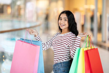 Foto de Joyful hermosa joven oriental mujer shopaholic mostrando sus compras, llevando bolsas de papel de colores en ambas manos y sonriendo a la cámara mientras camina por el centro comercial moderno - Imagen libre de derechos