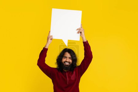 Téléchargez les photos : Portrait de joyeux jeune homme indien barbu souriant tenant blanc bulle de discours vide avec espace vide pour le texte sur fond jaune coloré studio - en image libre de droit