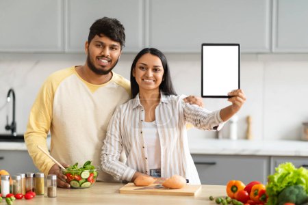 Foto de Blog de comida, recetas en línea. Positiva pareja india joven cocinar juntos en casa, mostrando tableta digital con pantalla en blanco espacio de copia maqueta para publicidad aplicación móvil, interior de la cocina moderna - Imagen libre de derechos