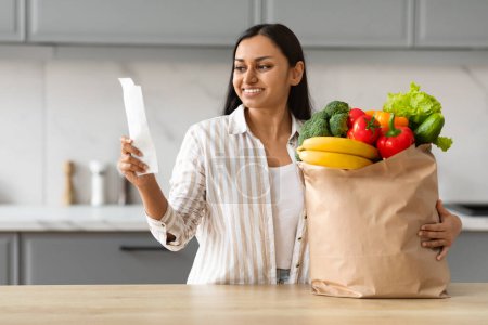 Photo for Happy pretty young indian woman with paper bag full of food fresh organic fruits and vegetable standing next to kitchen table, checking bill. Grocery delivery, economy shopping - Royalty Free Image