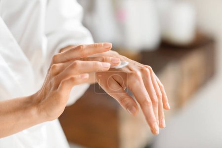 Cropped of young woman wearing white bathrobe enjoying her morning body care routine, applying cream lotion moisturizer on her hand after shower, bathroom interior, copy space