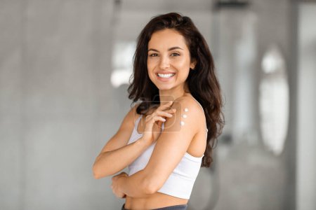 Photo for Home spa, body pampering concept. Smiling attractive brunette young woman with long hair wearing white top applying moisturizing body lotion on her shoulder after shower, copy space - Royalty Free Image