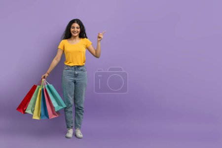 Cheerful woman holding shopping bags and pointing upwards with a smile on a purple background