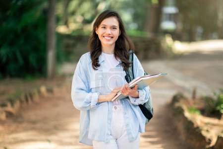 Estudiante joven europea feliz con una sonrisa encantadora sosteniendo cuadernos mientras camina con confianza en un parque, ejemplificando una vida de estudiantes en el campus, al aire libre. Estudio, educación