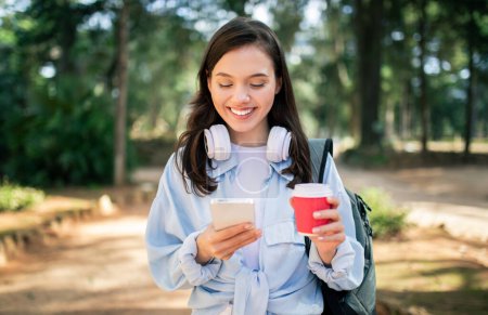 Joven estudiante europea contenta con auriculares revisando su teléfono inteligente y disfrutando de una bebida caliente en una taza roja desechable mientras camina en un parque, al aire libre. Chat, blog