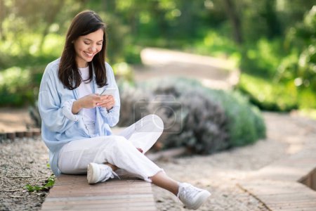 Téléchargez les photos : Contenu jeune étudiante européenne en vêtements décontractés textant sur son smartphone alors qu'elle était assise sur un sentier en bois dans un parc, entourée de verdure luxuriante en plein jour, en plein air - en image libre de droit