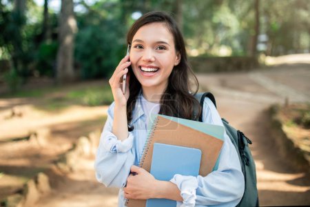 Joven estudiante europea alegre comprometida en una animada conversación telefónica mientras sostiene libros y una mochila, caminando en un entorno de parque cubierto de sol, al aire libre. Estudio, educación, llamada