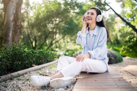 Téléchargez les photos : Une jeune étudiante européenne ravie avec des écouteurs blancs levant les yeux et souriant, profitant de la musique tout en étant assise sur un sentier en bois dans un parc serein, entouré de verdure - en image libre de droit