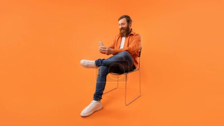 Téléchargez les photos : Homme d'affaires roux d'âge moyen avec la barbe défilant son smartphone avec le sourire, assis dans une chaise moderne sur fond de studio orange, mettant en valeur la technologie et la communication. Panorama - en image libre de droit