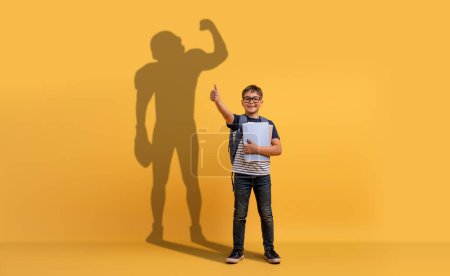 Téléchargez les photos : Un jeune garçon souriant avec des lunettes et un sac à dos tient des papiers scolaires, donnant un pouce devant un fond jaune avec une forte ombre de super-héros, représentant le potentiel et la confiance - en image libre de droit