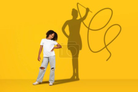 Téléchargez les photos : Une jeune fille confiante se tient avec sa main sur sa hanche dans des vêtements décontractés, jetant l'ombre d'une figure avec un lasso, représentant l'autonomisation et l'imagination créative sur un fond jaune - en image libre de droit