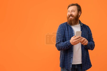 Téléchargez les photos : Heureux homme barbu aux cheveux roux en utilisant le téléphone et en regardant l'espace de copie, la publicité offre mobile ou nouvelle application, debout sur fond de studio orange. Gadgets et apps publicité - en image libre de droit