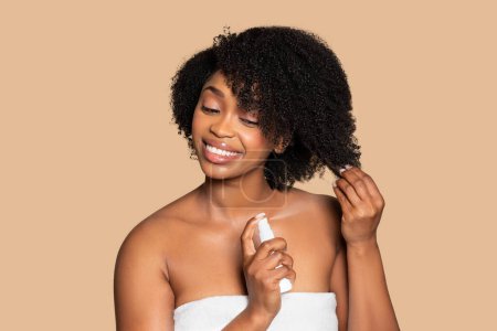 Foto de Joven y alegre mujer negra en toalla aplica un producto nutritivo para el cabello rizado, sonriendo agradablemente sobre un fondo beige cremoso - Imagen libre de derechos