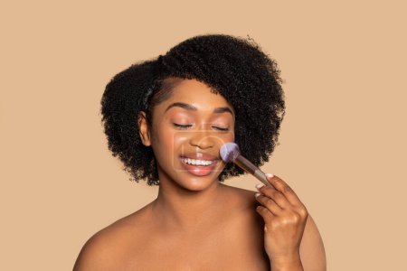 Foto de Mujer negra encantada con el pelo rizado aplicando rubor con cepillo, ojos cerrados en la felicidad, participando en su rutina de maquillaje contra el fondo neutro - Imagen libre de derechos