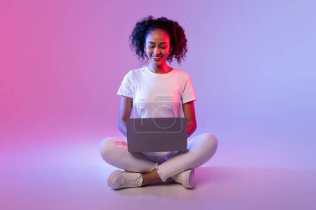 Eine fröhliche junge Frau genießt es, ihren Laptop zu benutzen, während sie auf dem Boden mit lila-rosa Hintergrund sitzt
