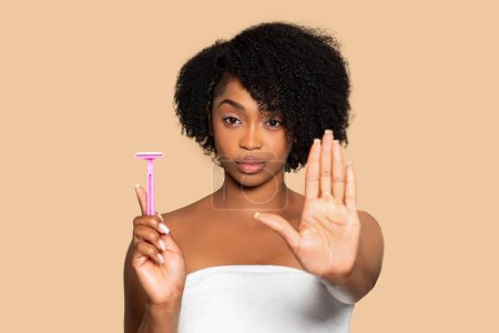 Téléchargez les photos : Femme noire habilitée avec une serviette, tenant un rasoir rose tout en faisant un geste d'arrêt, faisant un choix personnel au sujet de ses soins de la peau et de sa routine de cheveux du corps - en image libre de droit
