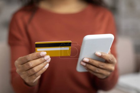 Photo for Young black woman holding credit card and using her smartphone at home, unrecognizable african american female making online payment or financial transaction, selective focus on hands - Royalty Free Image
