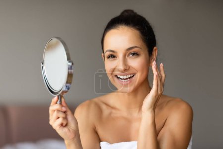 Téléchargez les photos : Jeune femme au teint éclatant parfait sourit à la caméra tout en tenant miroir rond, profiter d'une routine de soins de la peau saine et efficace dans la chambre moderne à la maison. Bien-être et beauté - en image libre de droit