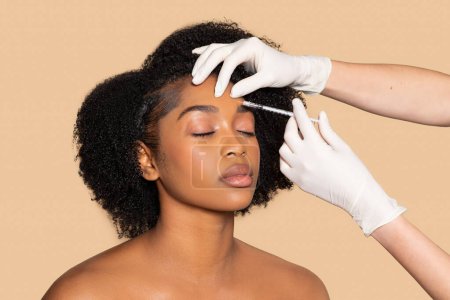 Téléchargez les photos : Femme noire sereine recevant un traitement méticuleux des sourcils, les yeux fermés en relaxation, car le cosmétologue applique un soin avec une seringue sur fond beige - en image libre de droit