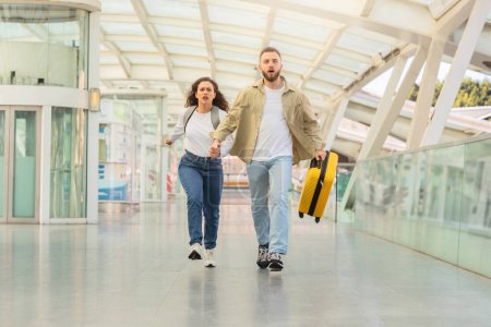 Determined couple sprinting through airport corridor with yellow suitcase, trying to catch flight, stressed man and woman holding hands while running with luggage at terminal, late to boarding