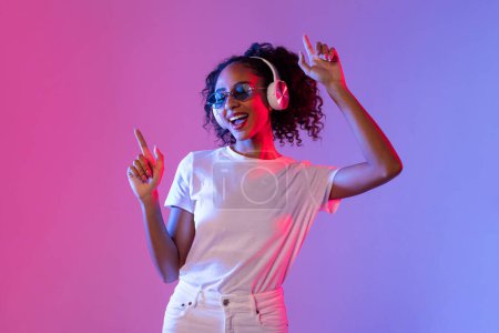 Foto de Mujer negra vibrante con el pelo rizado disfrutando de la música en los auriculares inalámbricos y bailando contra el fondo de neón degradado rosa y azul - Imagen libre de derechos