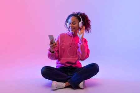Téléchargez les photos : Dame noire détendue en sweat à capuche rose assise jambes croisées, profitant de la musique avec écouteurs blancs tout en naviguant sur son smartphone en arrière-plan néon - en image libre de droit