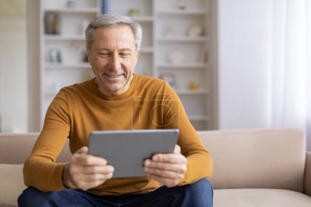 Photo for An elderly man is seen concentrating on a tablet while seated on a couch in a well-lit home environment - Royalty Free Image