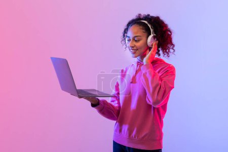 Photo for Smiling young woman in pink sweatshirt with headphones using an open laptop against neon lights - Royalty Free Image