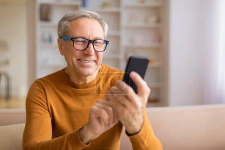 A smiling elder man with eyeglasses focused on his smartphone, conveying interest and comfort in a home environment