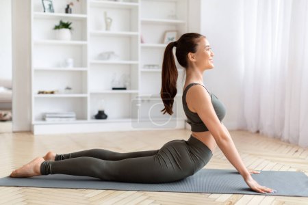 Téléchargez les photos : Belle jeune femme asiatique en vêtements de sport faisant posture cobra dos-étirement à la maison, attrayant entraînement coréen femme sur tapis de yoga dans lumineux, minimaliste style salon, vue latérale - en image libre de droit