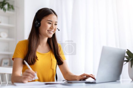 Foto de Sonriente joven coreana que usa auriculares mientras trabaja en su escritorio de oficina en casa ordenado y bien iluminado, mujer asiática feliz escribiendo en el portátil y tomando notas para una multitarea efectiva, espacio para copiar - Imagen libre de derechos