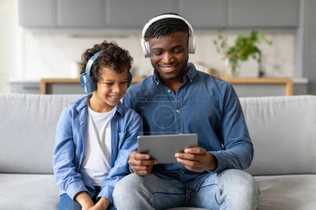 Téléchargez les photos : Père et fils vivent un moment joyeux en partageant du contenu sur une tablette numérique avec des écouteurs - en image libre de droit
