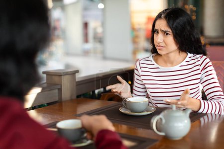Foto de Irritada joven mujer india atractiva mirando a su novio con duda, pareja oriental enojado sentado en la cafetería. Los cónyuges orientales se pelean durante la cita en la cafetería - Imagen libre de derechos