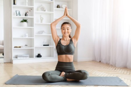 Foto de Mujer asiática joven pacífica en ropa deportiva practicando yoga en casa, hermosa hembra coreana con sonrisa serena, sentada en pose de loto en la colchoneta de fitness en el interior de la sala de estar bien iluminada, espacio para copiar - Imagen libre de derechos