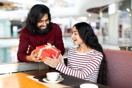 Foto de Un indio cariñoso dando a su novia un regalo de cumpleaños en la cafetería. Romántica pareja oriental joven celebrando aniversario juntos en la cafetería, intercambiando regalos entre sí - Imagen libre de derechos