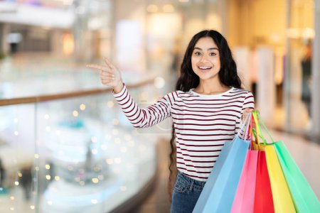 Photo for Positive pretty long-haired young indian woman carrying colorful paper bags purchases, walking by modern city center, pointing at copy space. Black friday, season sale concept - Royalty Free Image