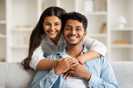 Photo for Happy indian couple embracing on couch, with the woman behind the man, both smiling at camera, radiating love and happiness in comfortable home setting - Royalty Free Image