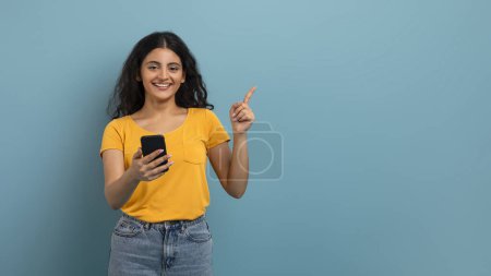 Zufriedene Frau, die ihr Smartphone benutzt und mit einem netten Lächeln auf blauem Hintergrund nach links zeigt