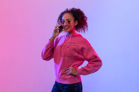 Foto de Mujer negra de moda con el pelo rizado hablando en el teléfono inteligente, con gafas de sol redondas y sudadera con capucha rosa, con un vibrante fondo rosa neón y azul - Imagen libre de derechos