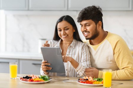 Téléchargez les photos : Conjoints indiens joyeux utilisant la tablette numérique tout en mangeant le petit déjeuner dans la cuisine, heureux couple oriental appréciant savoureux repas du matin, shopping en ligne ou la vérification des réseaux sociaux sur Pad, espace libre - en image libre de droit