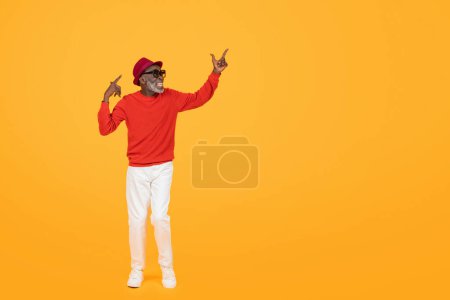 Téléchargez les photos : Élégant homme noir âgé heureux dans un pull rouge et chapeau, avec des lunettes de soleil, pointant vers le haut avec les deux mains, dansant et s'amusant sur un fond jaune, studio, pleine longueur - en image libre de droit
