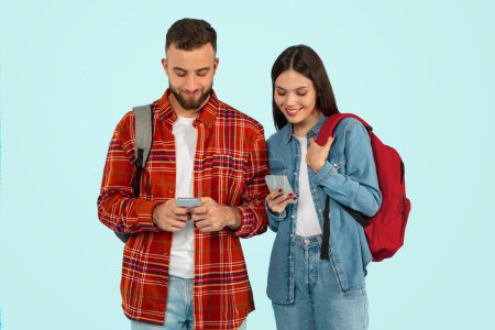 Photo for Young couple college students with backpacks immersed in social media on their smartphones, symbolizing modern communication and youth connection, against blue studio backdrop - Royalty Free Image