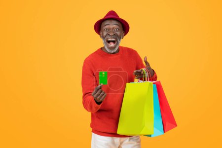 Téléchargez les photos : Joyeux homme noir âgé dans un chapeau rouge vibrant et pull, tenant une carte de crédit et des sacs à provisions colorés avec un sourire à la bouche grande ouverte sur un fond orange vif, studio - en image libre de droit
