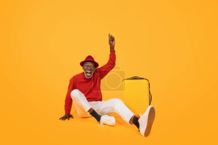 Foto de Hombre negro mayor alegre en un suéter rojo y sombrero sentado en el suelo con un gesto de victoria levantado, junto a una maleta amarilla sobre un fondo naranja vibrante, estudio - Imagen libre de derechos