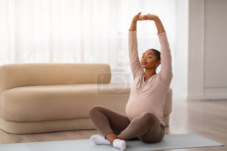 Téléchargez les photos : Paisible jeune femme noire enceinte ont yoga à la maison, assis sur tapis de fitness avec les yeux fermés au salon ensoleillé, levant les mains vers le haut. Entraînement pendant la grossesse, espace de copie - en image libre de droit