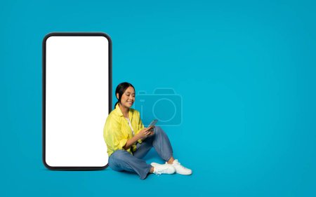 Photo for Smiling millennial Asian woman in yellow sits cross-legged, enjoying her smartphone beside a giant smartphone mockup with a blank screen on a bright turquoise background, studio - Royalty Free Image